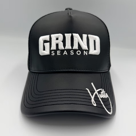 A black structured trucker-style cap with white embroidered text reading 'GRIND SEASON' on the front and a white script signature on the brim, photographed against a light gray studio background.