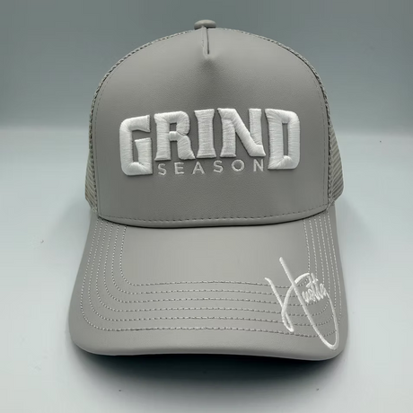A gray trucker-style baseball cap with white embroidered text reading 'GRIND SEASON' on the front and a white script signature on the brim, shown against a light gray studio background.