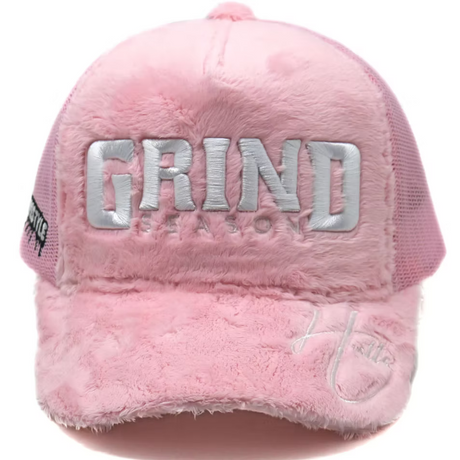 A pink fuzzy trucker hat with 'GRIND' and 'SEASON' embroidered in silver on the front, featuring a pink mesh back and a small script logo on the brim.