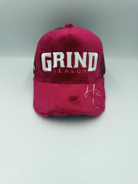 A fuzzy, plush trucker-style cap in a deep magenta pink color, featuring 'GRIND SEASON' embroidered in white on the front. The cap has a dark mesh back and a small white script logo on the brim, set against a neutral gray studio background.