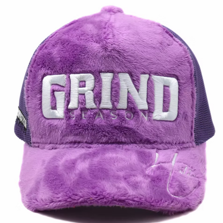 A purple fuzzy trucker cap with dark purple mesh side panels and large silver embroidered text reading 'GRIND' and 'SEASONS' on the front.